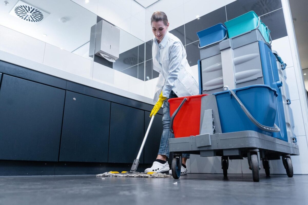 A healthcare facility cleaner mopping the floor in a restroom with a large cleaning cart beside her.